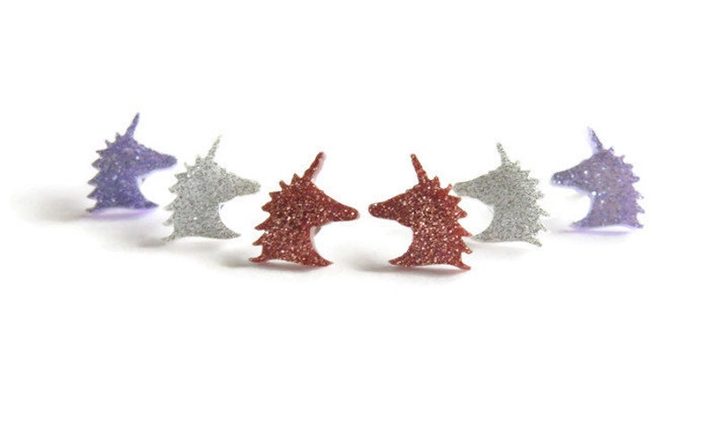 May include: Glittery unicorn head earrings in purple, silver, and red-orange. The earrings are arranged in a semi-circle, each with a horn and detailed mane. The earrings are small, likely stud earrings, and are set against a white background.