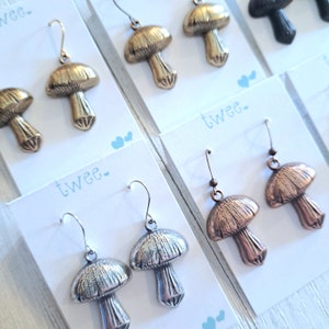 Close up of several pairs of mushroom earrings on white and aqua earring cards. Detail is shown on antiqued brass with gold wires, antiqued silver, and antiqued copper, versions.