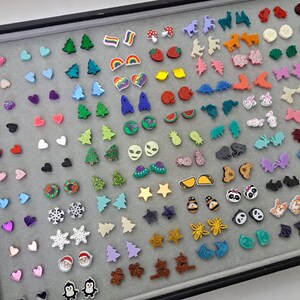 May include: A collection of colorful stud earrings in various shapes, including hearts, stars, Christmas trees, and animals. The earrings are displayed on a gray surface, showcasing a variety of designs and colors, perfect for adding a touch of fun to any outfit.