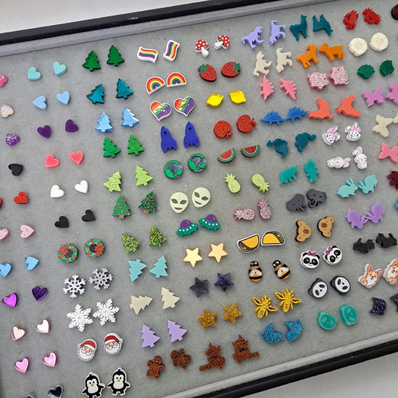 May include: A collection of colorful stud earrings in various shapes, including hearts, stars, Christmas trees, and animals. The earrings are displayed on a gray surface, showcasing a variety of designs and colors, perfect for adding a touch of fun to any outfit.