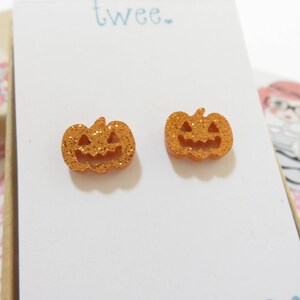 Puede incluir: Un par de pendientes de calabaza naranja con purpurina. Los pendientes tienen forma de calabaza con una cara sonriente.
