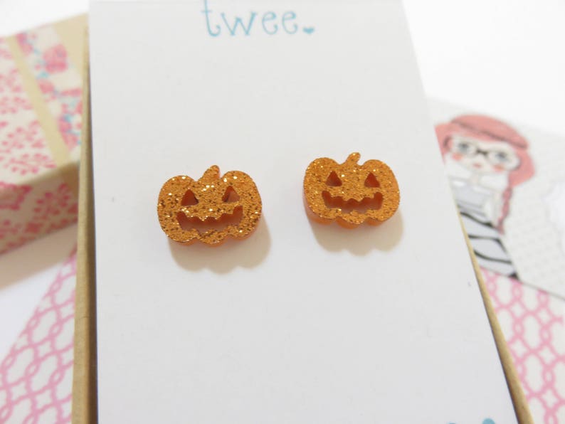 Puede incluir: Un par de pendientes de calabaza naranja con purpurina. Los pendientes tienen forma de calabaza con una cara sonriente.