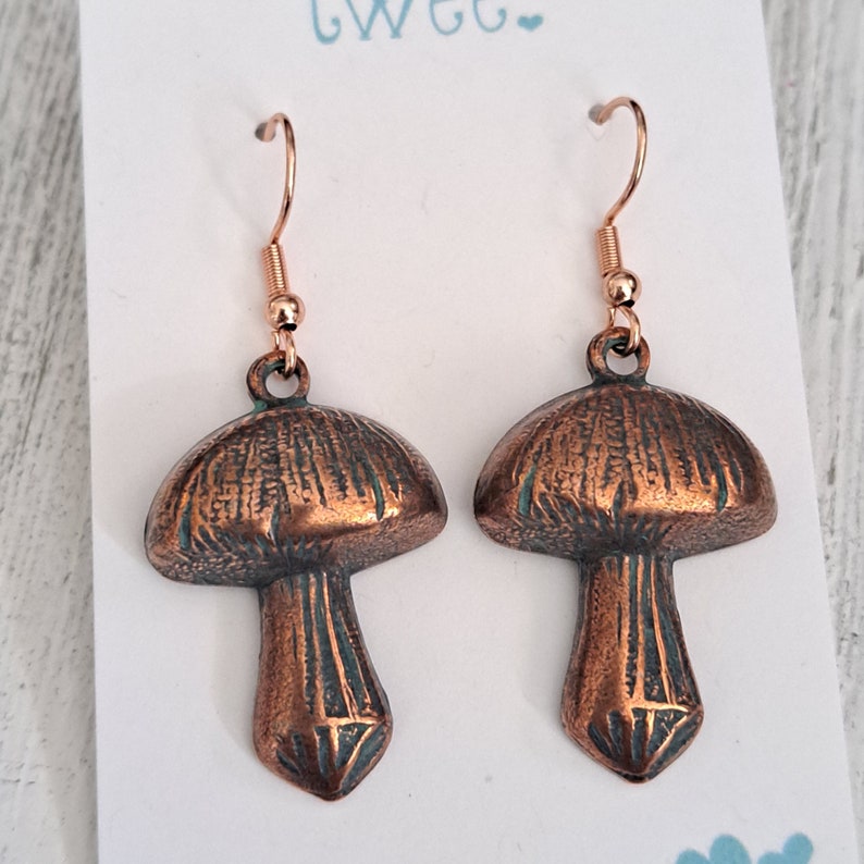 Close up of mushroom earrings in copper with a verdigris patina and bright copper ear wires. Shown on a white earring card with aqua colored Twee at the top and has 2 little hearts in the lower right corner.