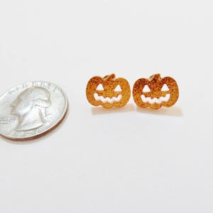 Puede incluir: Un par de pendientes de calabaza de purpurina dorada con caras de calabaza blancas.