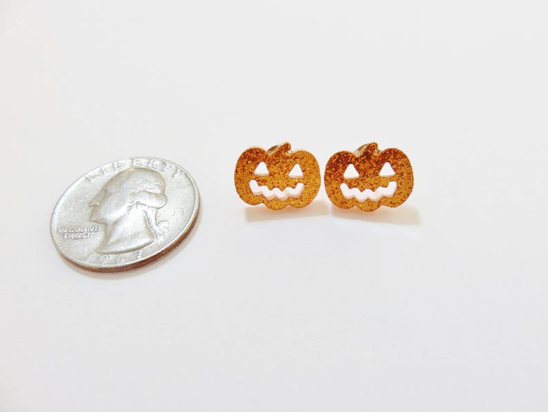 Puede incluir: Un par de pendientes de calabaza de purpurina dorada con caras de calabaza blancas.