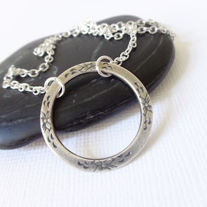 Puede incluir: Un collar con colgante plateado con un colgante circular adornado con grabados florales. El colgante cuelga de una fina cadena. El collar descansa sobre una piedra oscura y lisa sobre un fondo blanco, resaltando el diseño de la joya.