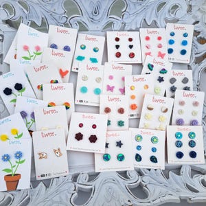 May include: A collection of stud earrings on white cards, each featuring the word "twee." Designs include flowers, animals, hearts, stars, and geometric shapes in various colors. The earrings are displayed on a decorative white surface.