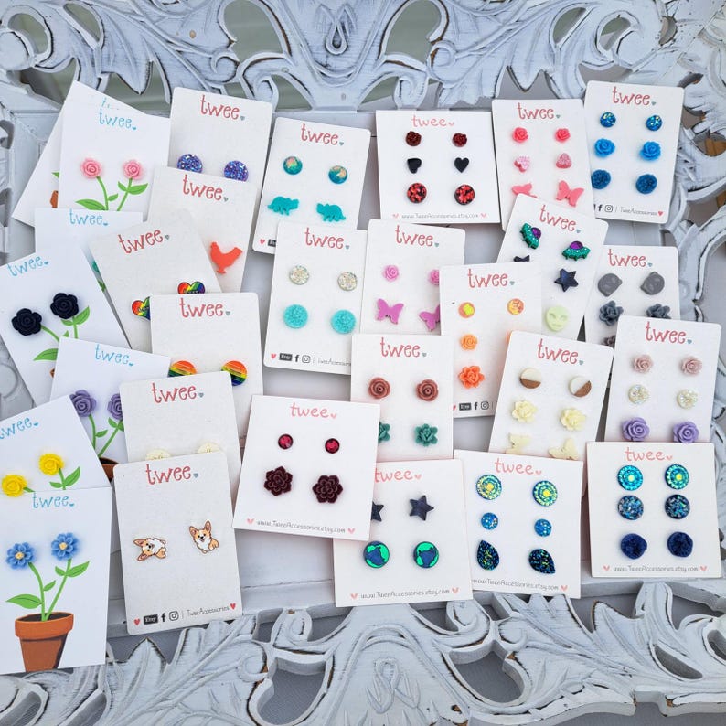 May include: A collection of stud earrings on white cards, each featuring the word "twee." Designs include flowers, animals, hearts, stars, and geometric shapes in various colors. The earrings are displayed on a decorative white surface.