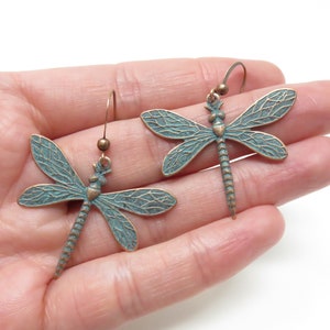 May include: A pair of dangle earrings featuring a dragonfly design. The dragonflies are made of metal with a green patina finish and have detailed wings. The earrings are suspended from a simple hook.