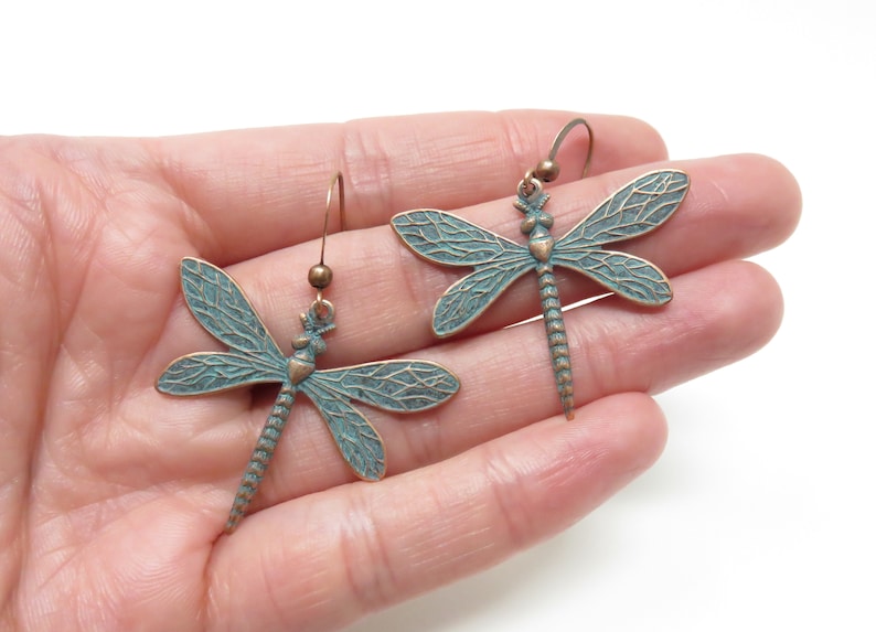 May include: A pair of dangle earrings featuring a dragonfly design. The dragonflies are made of metal with a green patina finish and have detailed wings. The earrings are suspended from a simple hook.