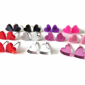 May include: A collection of heart-shaped stud earrings in various colors, including red, pink, purple, black, and clear. Some earrings have glitter or a mirrored finish. The earrings are displayed on a white background.