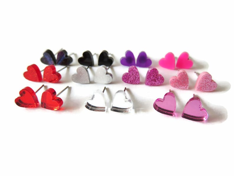 May include: A collection of heart-shaped stud earrings in various colors, including red, pink, purple, black, and clear. Some earrings have glitter or a mirrored finish. The earrings are displayed on a white background.