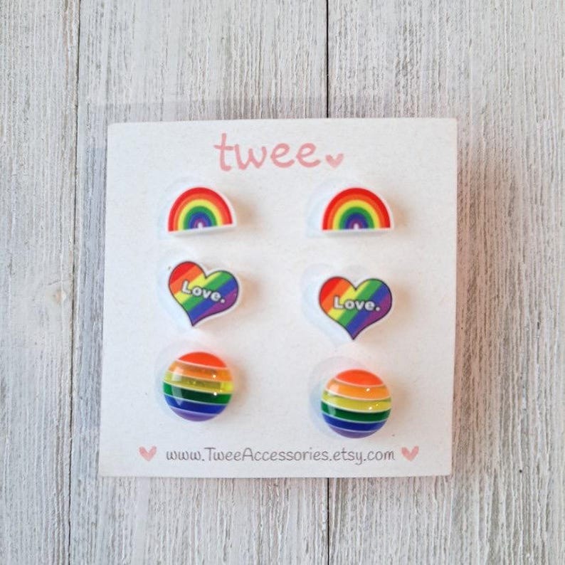 May include: A set of six colorful Pride-themed earrings on a white card. The earrings feature rainbow arches, hearts with "Love" text, and striped spheres. The card has the word "twee" and a website address printed on it.