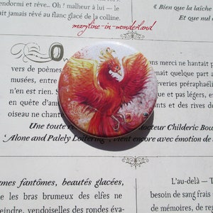 Può includere: Un bottone rotondo con un vivace disegno di fenice nei toni del rosso, arancione e giallo. Il bottone ha uno sfondo bianco e un bordo metallico. Il testo "maryline-in-wonderland" è visibile sullo sfondo.