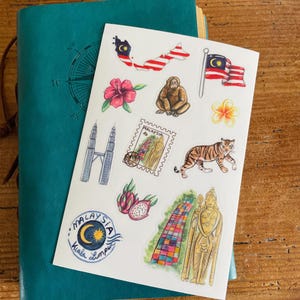 Puede incluir: Una hoja de pegatinas con temática de Malasia, con ilustraciones de la bandera, el mapa, flores, animales y monumentos. Las pegatinas están sobre un fondo blanco, apoyadas en un diario turquesa. También se ve el emblema de Kuala Lumpur.