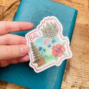 May include: A colorful sticker with the word "BALI" features a Balinese temple, a red hibiscus flower, and a traditional mask. The sticker is held over a teal notebook with a compass rose design on a wooden surface.