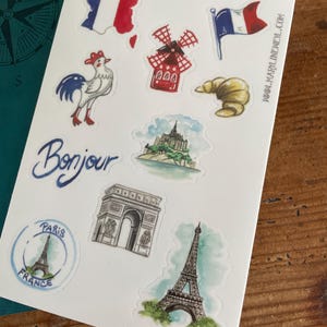 May include: A sheet of stickers featuring French-themed illustrations. Designs include a map of France, a rooster, a windmill, a croissant, a flag, the Eiffel Tower, the Arc de Triomphe, and the word "Bonjour". The stickers are on a white background.
