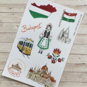 May include: A sticker sheet featuring illustrations of Budapest landmarks and symbols, including the Hungarian flag, a yellow tram, a person in traditional attire, a floral motif, the Parliament building, and the text Budapest and www.marylineweyl.com.