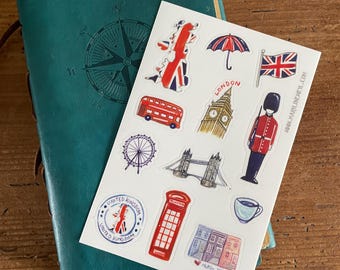 Op de afbeelding: Een turquoise leren notitieboek met een kompasroosontwerp ligt naast een stickervel met Londense bezienswaardigheden. De stickers tonen een rode dubbeldekkerbus, Big Ben en een Britse vlag.
