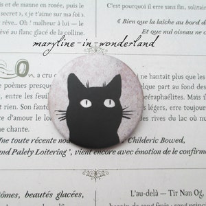 Puede incluir: Un botón redondo con una silueta de gato negro con ojos y bigotes blancos sobre un fondo lila claro. El botón tiene el texto "maryline-in-wonderland" en cursiva. Un accesorio con temática de gatos.