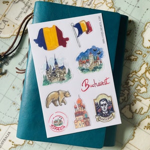 Op de afbeelding: Een vel met kleurrijke stickers met Roemeense symbolen, waaronder de vlag, een beer en kastelen. Het woord "Bucharest" staat in rode letters. De stickers liggen op een wit vel, op een blauwgroen notitieboek.