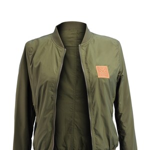 May include: A green bomber jacket with a zipper closure and a brown leather patch with a logo on the right side of the chest.