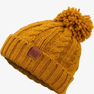 Könnte beinhalten: Eine senfgelbe Strickmütze mit einem braunen Lederflicken und einem flauschigen Bommel oben.