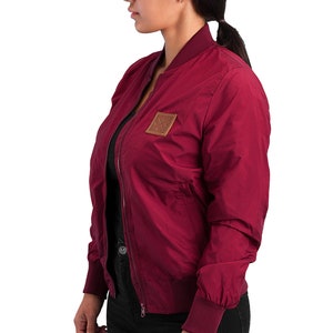 May include: A maroon bomber jacket with a brown leather patch on the left side. The jacket has a zipper closure and ribbed cuffs and waistband.