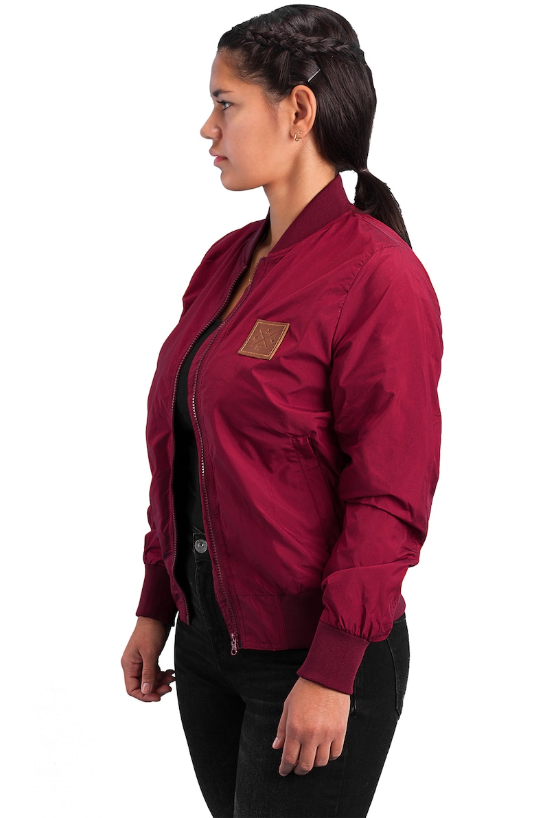 May include: A maroon bomber jacket with a brown leather patch on the left side. The jacket has a zipper closure and ribbed cuffs and waistband.