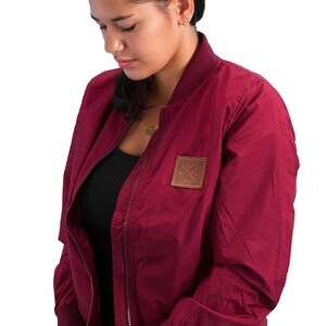 May include: A maroon bomber jacket with a zipper closure and a brown patch on the left chest with a logo of crossed wrenches and the text "1981".