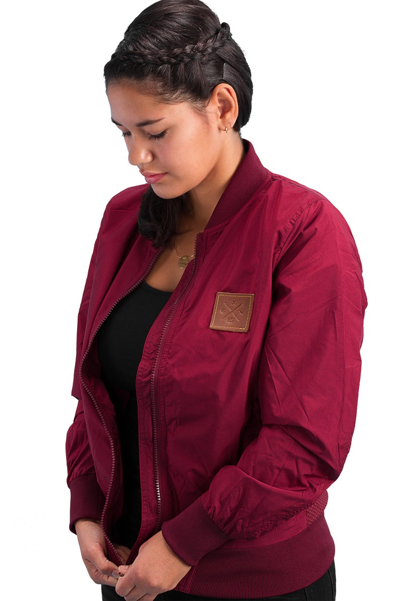 May include: A maroon bomber jacket with a zipper closure and a brown patch on the left chest with a logo of crossed wrenches and the text "1981".