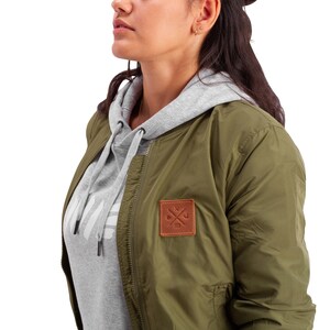 May include: A green bomber jacket with a brown leather patch featuring a crossed wrench and anchor design. The jacket has a zipper closure and a gray hooded sweatshirt underneath.