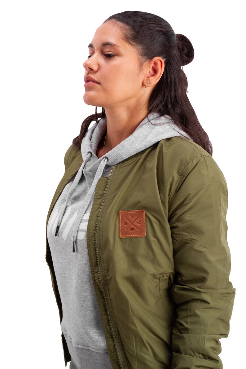 May include: A green bomber jacket with a brown leather patch featuring a crossed wrench and anchor design. The jacket has a zipper closure and a gray hooded sweatshirt underneath.