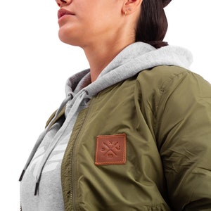 May include: A green bomber jacket with a brown leather patch featuring a compass and the text "XIX". The jacket has a zipper closure and a gray hooded sweatshirt underneath.