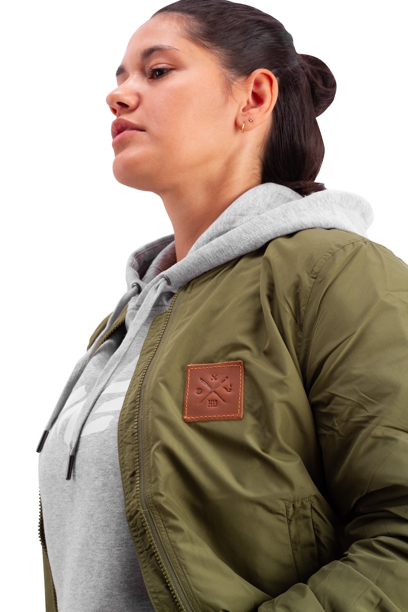 May include: A green bomber jacket with a brown leather patch featuring a compass and the text "XIX". The jacket has a zipper closure and a gray hooded sweatshirt underneath.