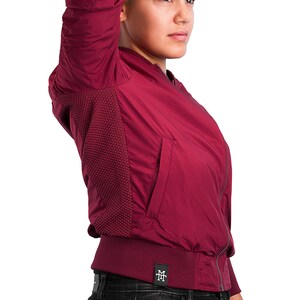 May include: A burgundy bomber jacket with a zipper closure and a small logo patch on the bottom left side. The jacket has a ribbed collar and cuffs.