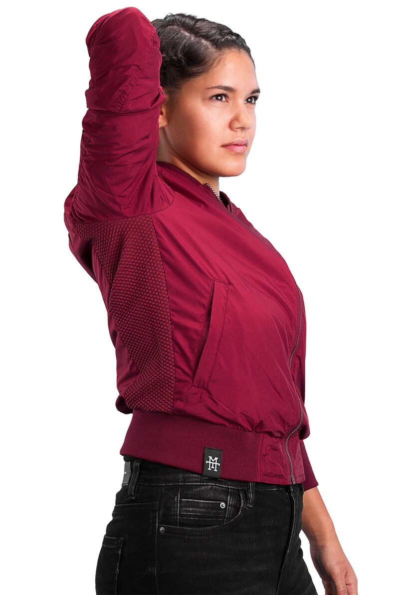 May include: A burgundy bomber jacket with a zipper closure and a small logo patch on the bottom left side. The jacket has a ribbed collar and cuffs.