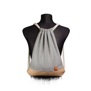 Könnte beinhalten: Ein grauer Rucksack mit Kordelzug und einem braunen Lederpatch. Der Rucksack ist aus weichem, bequemem Stoff gefertigt und hat ein schlichtes, elegantes Design.