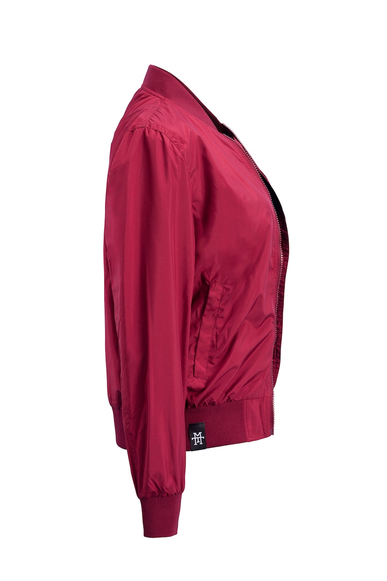 May include: A burgundy bomber jacket with a zipper closure and a small logo on the left side. The jacket has a ribbed collar, cuffs, and hem.