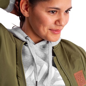 May include: A woman wearing a green bomber jacket with a gray hoodie underneath. The jacket has a brown patch with a white cross on it. The hoodie has a white graphic on the front.