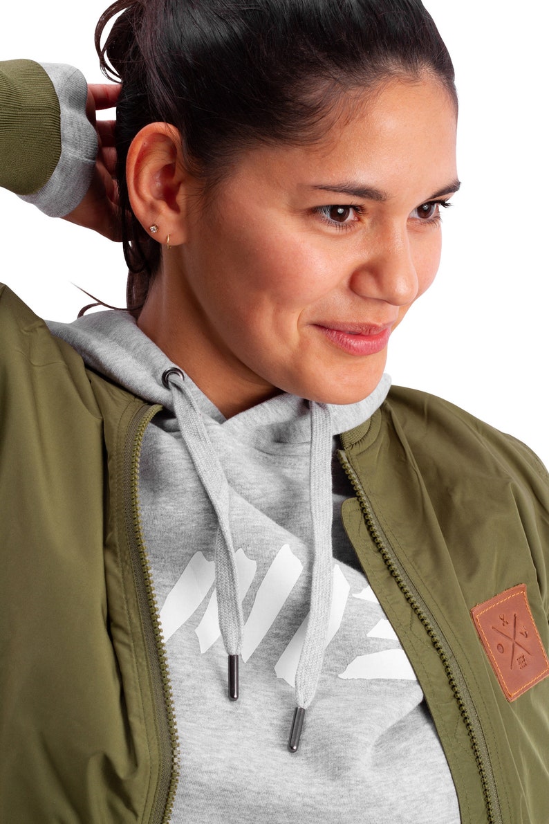 May include: A woman wearing a green bomber jacket with a gray hoodie underneath. The jacket has a brown patch with a white cross on it. The hoodie has a white graphic on the front.