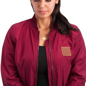 May include: A maroon bomber jacket with a brown leather patch on the left side featuring a cross and the text "1973".