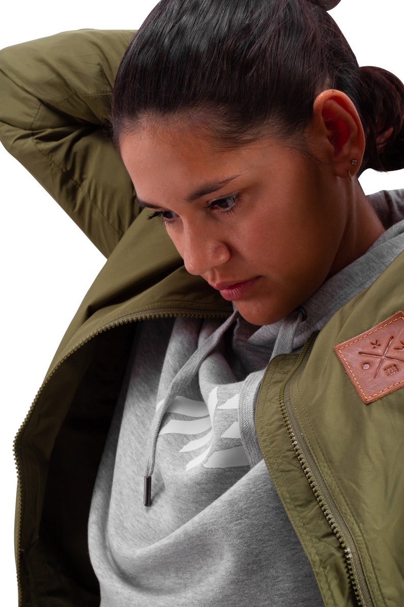 May include: A woman wearing a green bomber jacket with a zipper and a brown patch with a logo. She is also wearing a grey hoodie with a white logo.