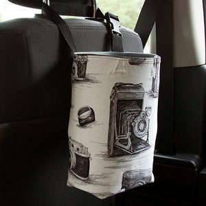 May include: A car trash bag with a black strap and buckle, hanging from a car headrest. The bag is white with a black and gray vintage camera pattern. The bag is rectangular and hangs vertically.