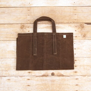 Pu&ograve; includere: Una borsa tote in tela marrone con due manici. La borsa ha una piccola etichetta verde sul davanti.