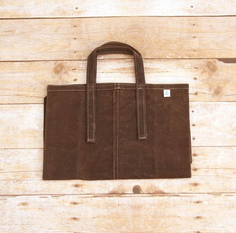 Pu&ograve; includere: Una borsa tote in tela marrone con due manici. La borsa ha una piccola etichetta verde sul davanti.
