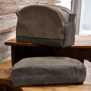 May include: Two canvas toiletry bags in shades of olive green and charcoal gray. The top bag is a rounded shape with a handle, and the bottom bag is rectangular. Both bags are sitting on a wooden surface.