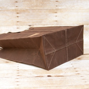 Pu&ograve; includere: Una borsa tote in tela marrone con un fondo rinforzato e un'unica maniglia. La borsa &egrave; piegata e appoggiata su una superficie di legno.