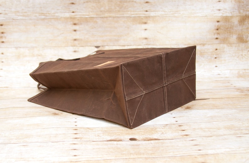 Pu&ograve; includere: Una borsa tote in tela marrone con un fondo rinforzato e un'unica maniglia. La borsa &egrave; piegata e appoggiata su una superficie di legno.