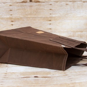 Pu&ograve; includere: Una borsa tote in tela marrone con una toppa in pelle e due manici. La borsa &egrave; piegata e appoggiata su una superficie di legno.